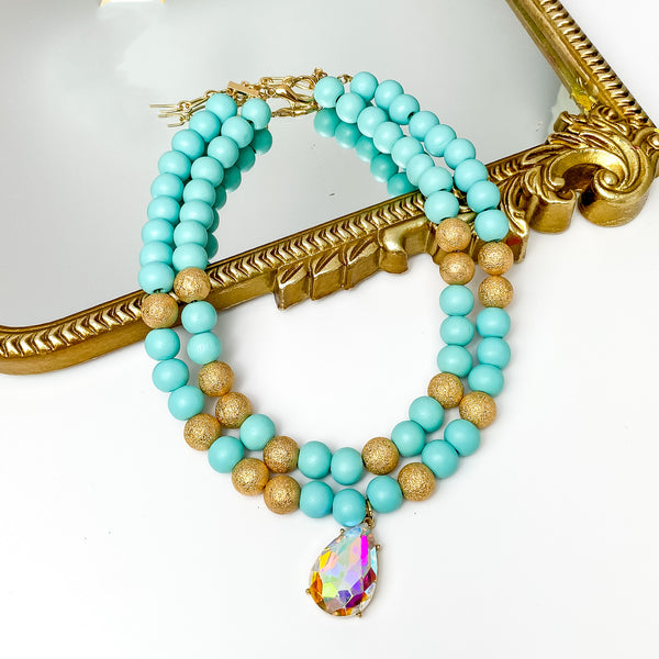 Two strand beaded necklace with a large ab crystal teardrop pendant. This necklace includes turquoise and gold beads. This necklace is pictured partially laying on a gold mirror on a white background. 
