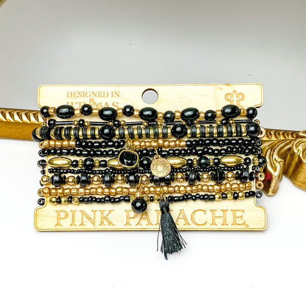 Set of ten black and gold bead bracelets in different sizes.These bracelets also include a black cushion cut crystal, a gold circle charm, a black bead charm, and a black tassel charm. These braceets are pictured on a Pink Panache wood holder in front of a gold mirror and on a white background. 