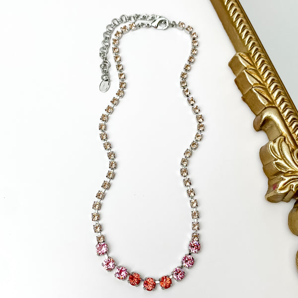 Pictured is a silver necklace with a mix of pink crystals. This necklace is pictured on a white background with a gold mirror on the right side.