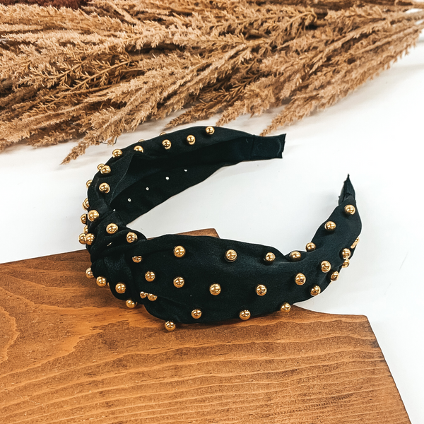 Black headband that is a knot in the middle of the headband. There are also gold beads throughout the entire headband. This headband is pictured laying in a piece of wood with tan floral at the top of the picture on a white background.