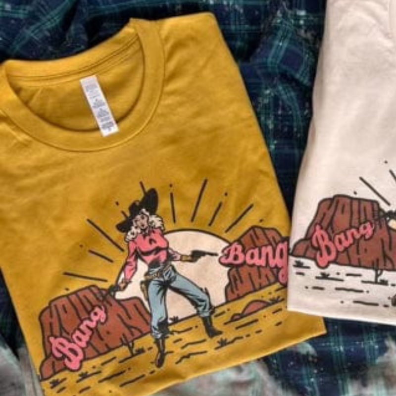 This mustard yellow graphic tee includes a crew neckline, short sleeves, and cute hand drawn design of a western scene of a cowgirl in front of mountains and the sun with pistols and the words "Bang Bang" in pink bubble font on either side of her. This tee is shown in this photo as a folded flat lay.