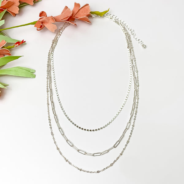 Silver tone three layered chain necklace. Pictured on a white background with flowers at the top left.