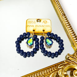 Circle drop navy beaded earrings with a hanging black ab teardrop crystal. These earrings are pictured on a wood earrings holder on a gold mirror on a white background. 
