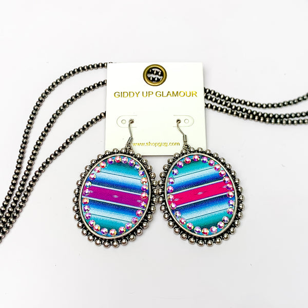 These earrings are oval shaped with aztec print inlay with the outline of the earing is silver but also has ab crystal going around the oval shape. These pair of earrings are pictured on a white background with navajo pearls behind it.