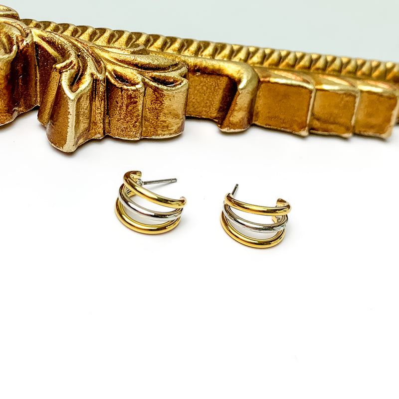 A pair of triple hoop earrings with two gold hoops and one silver hoop in the middle. These earrings are pictured on a white background with a gold material at the top of the picture. 