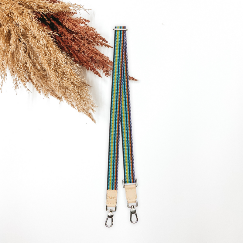 A woven multicolored purse strap that has a main color of black. This purse strap is pictured on a white background with tan and brown pompous grass in the top left corner.