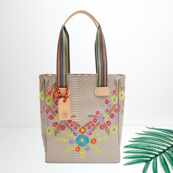 Centered in the picture is a cream colored tote with multi colored embroidery, To the right of the tote is a palm leaf, all on a white background. 