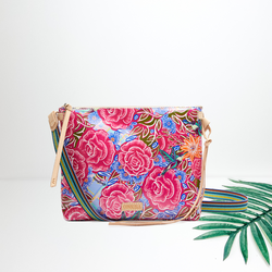 A pink floral square purse with a multicolor crossbody strap and leather detailing. Pictured on white background with a straw hat and palm leaf.