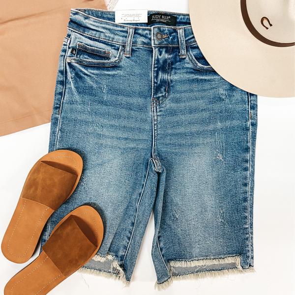 A pair of bermuda shorts that have a cut-off hemline and a light wash. Pictured with brown tank top, a pair of tan sandals, and a silverbelly hat.