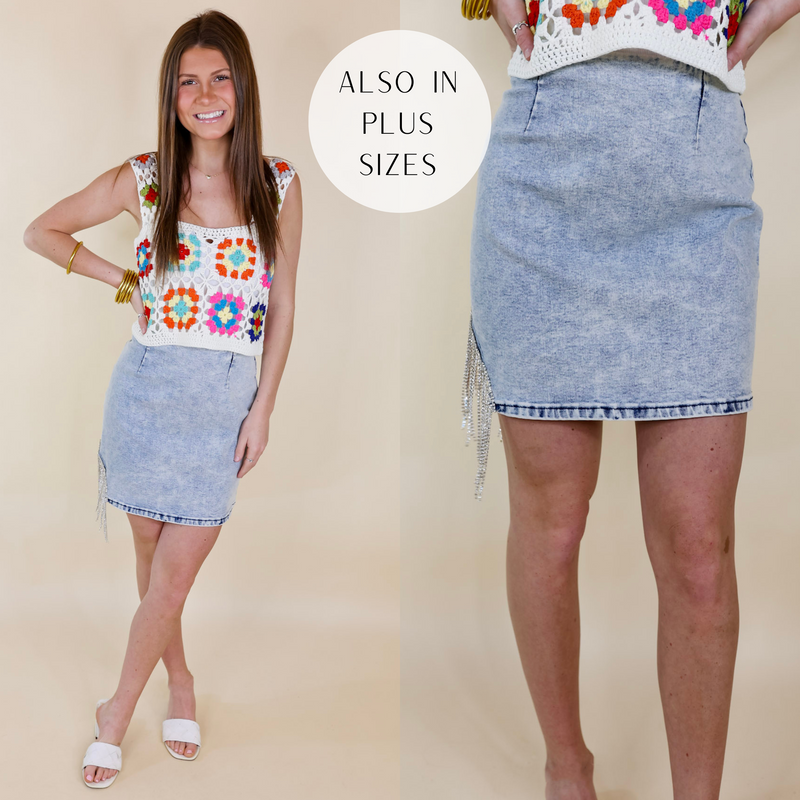 Model is wearing a light wash denim skirt with crystal fringe with crystal fringe on the hemline. Model has this top paired with white heels, a crochet tank top, and gold jewelry.