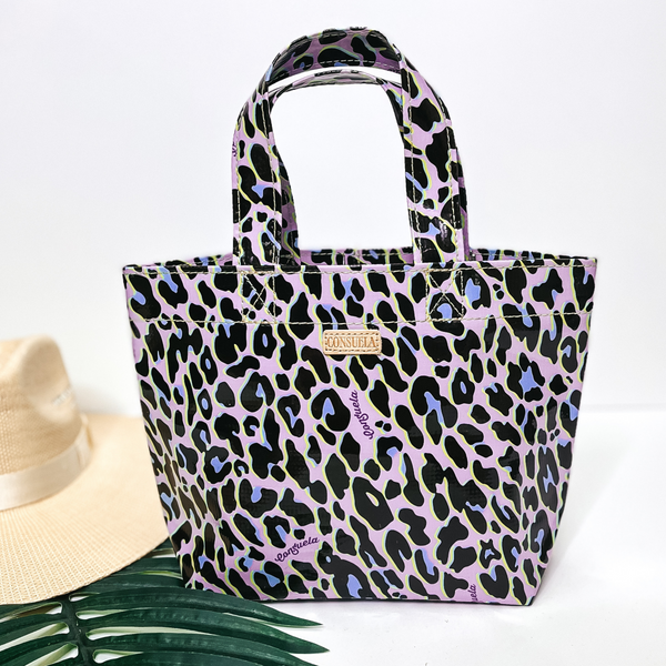 A small size purple  leopard print bag with handles. Pictured on white background with palm leaf and straw hat.