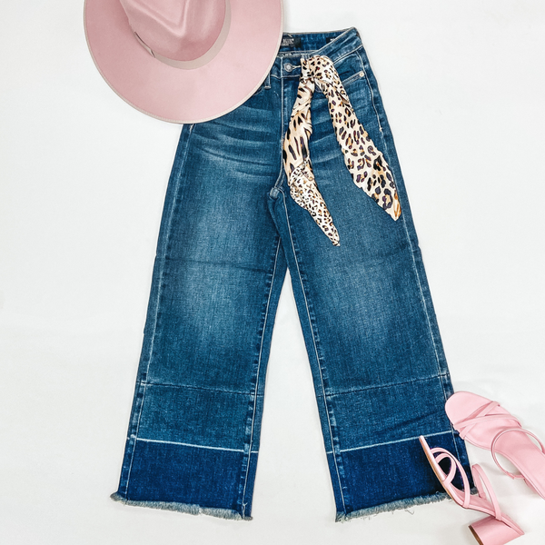 A pair of cropped wide leg jeans with a dark wash and raw hem. Pictured on a white background with pink heels, a pink hat, and a leopard print wild rag.