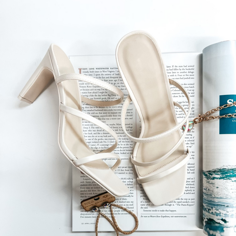 A pair of ivory strappy heels that have a square toe. These shoes are pictured on a white background with gold jewelry and a magazine.
