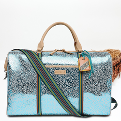 Metallic blue leopard duffle bag with tan leather handles. This bag also has a striped strap. This pictured on a white background with tan and brown pompous grass in the top left corner. 