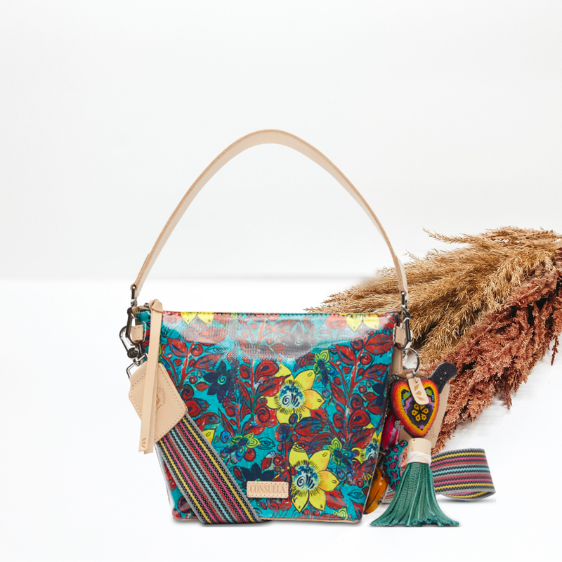 In the middle of the picture is a wedge bag in a blue, yellow, and red floral print. The background is solid white with pompas grass to the right of the bag.