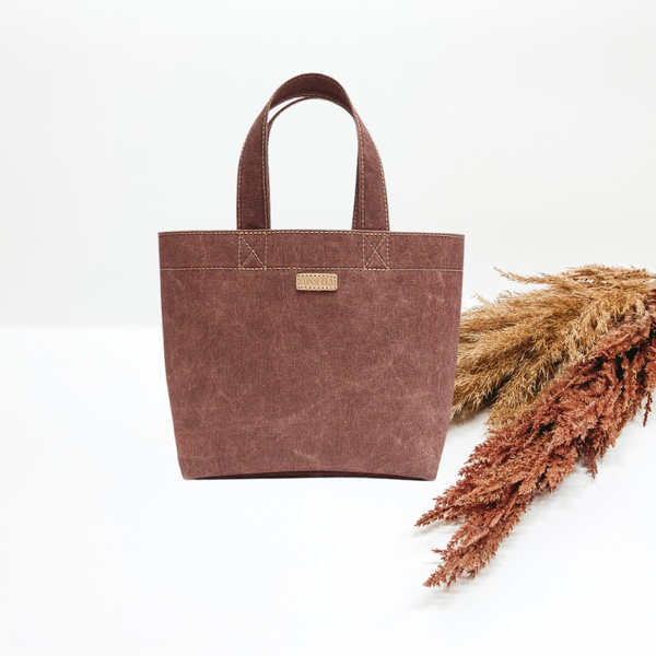 Pictured is a small tote bag in a brown-mauve color. Background is solid white with pompas grass laying to the right of the bag.  