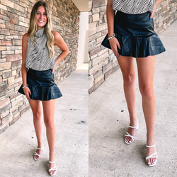 Model is wearing a black faux leather skirt with a ruffle hem. Model has it paired with a striped top, white heels, and crystal jewelry.