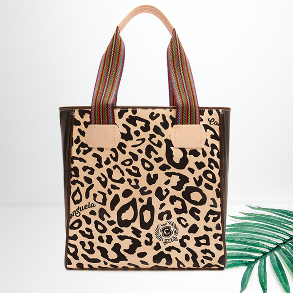 Centered in the picture is a tote bag in a tan cheetah pattern. To the right of the tote is a palm leaf on a white background. 