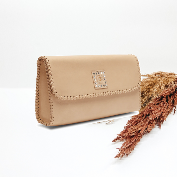 Centered in the picture is a natural colored clutch bag with rhinestones on the Consuela emblem. Background is solid white with pompas grass on the right side.  