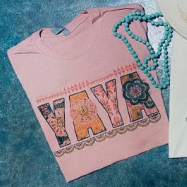 A light pink short sleeve tee with the word "YAYA" in the center. Each letter has its own unique pattern and color. There is a scalloped border below the text and a line border above. Item is pictured on a simple turquoise background.