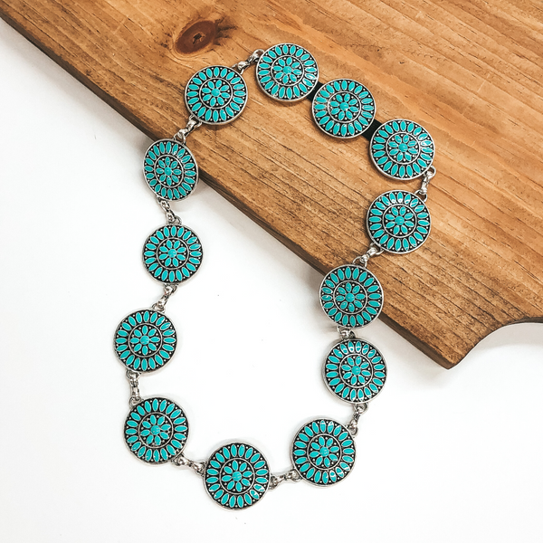 Silver, circle concho hat band with turquoise stones. This hat band is pictured partially laying on a brown block on a white background.