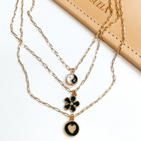 Three stranded gold chain necklace. Each strand has an individual charm in black, white, and/or gold. These include, a ying and yang circle charm, a flower charm, and a circle charm with a gold heart. This necklace is pictured partially on a tan bag on a white background. 