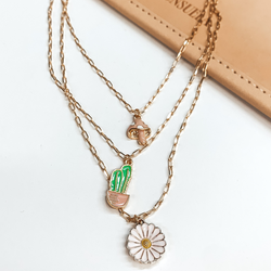 Three stranded gold chain necklace. Each strand has an individual charm in colors white, green, white, and pale pink. These include, a mushroom charm, a cactus in a pot charm, and a white flower charm. This necklace is pictured partially on a tan bag on a white background. 