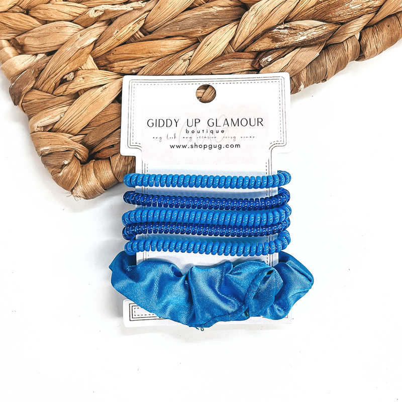 A set of six hair ties, five spiral and one satin scrunchie in blue. There are three  solid color spiral hair ties and two transparent. These hair ties are on a white  cardboard piece with a thekitchenapproach logo, they are leaned up against a brown woven  plate and a white background.