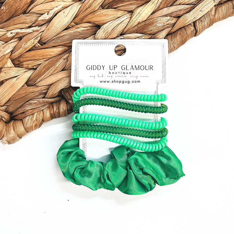 A set of six hair ties, five spiral and one satin scrunchie in green. There are three  solid color spiral hair ties and two transparent. These hair ties are on a white  cardboard piece with a thekitchenapproach logo, they are leaned up against a brown woven  plate and a white background.