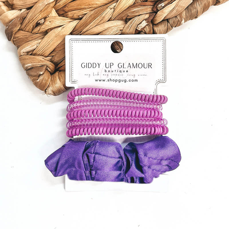 A set of six hair ties, five spiral and one satin scrunchie in purple. There are three  solid color spiral hair ties and two transparent. These hair ties are on a white  cardboard piece with a thekitchenapproach logo, they are leaned up against a brown woven  plate and a white background.