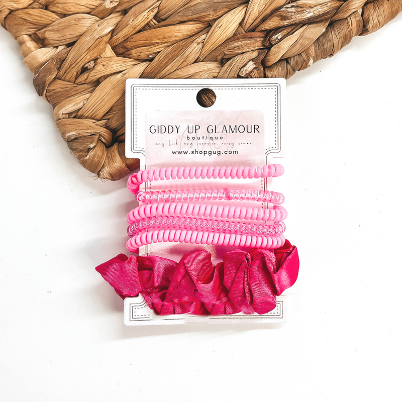 A set of six hair ties, five spiral and one satin scrunchie in hot pink. There are three  solid color spiral hair ties and two transparent. These hair ties are on a white  cardboard piece with a thekitchenapproach logo, they are leaned up against a brown woven  plate and a white background.