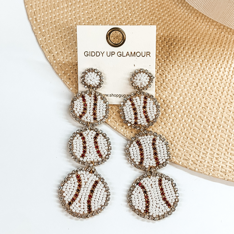 White beaded post back earrings with three beaded baseball pendants. These earrings also include a black crystal outline. These earrings are pictured partially laying on a straw hat brim on a white background. 