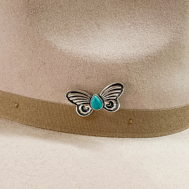 Silver butterfly hat pin with a center, teardrop, turquoise stone pictured on a beige colored hat pictured on a white background.