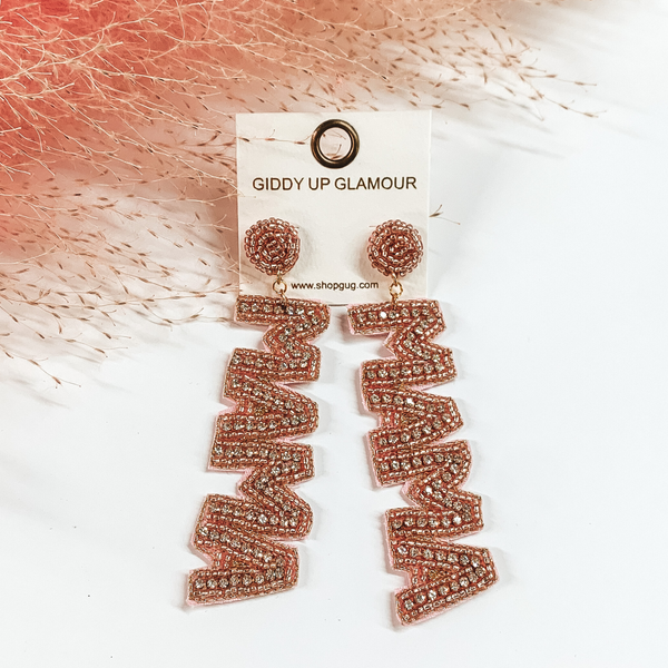 These earrings include rose pink, circle, beaded post back earrings with a long dangle. This dangle spells out "MAMA" in rose pink beads with an inlay of clear crystals. These earrings are pictured on a white background with pink pompous grass at the top. 