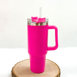 This is a hot pink tumbler with a hot pink handle. This tumbler also has a clear lid and straw. This tumbler is pictured standing on a piece of wood on a white background. 
