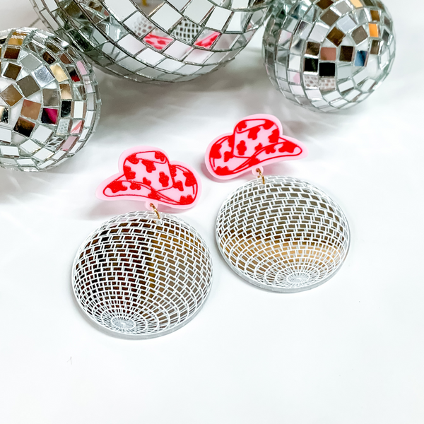 Pink cowprint cowboy hat earrings with a disco ball drop. These earring are pictured on a white background with disco balls at the top of the picture. 