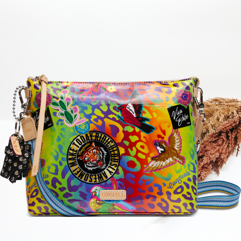 Pictured is a crossbody purse with a mostly black multicolored leopard print design. This purse also includes random stickers all over the leopard print. This purse is pictured on a white background with pompous grass on the right side of the picture.