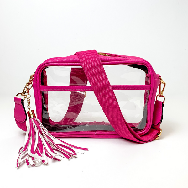 Pictured on a white background is a clear, rectangle purse with a fuchsia pink outline. This purse includes gold accessories, a fuchsia pink strap that is draped over the purse, and a pink and white fringe tassel. 