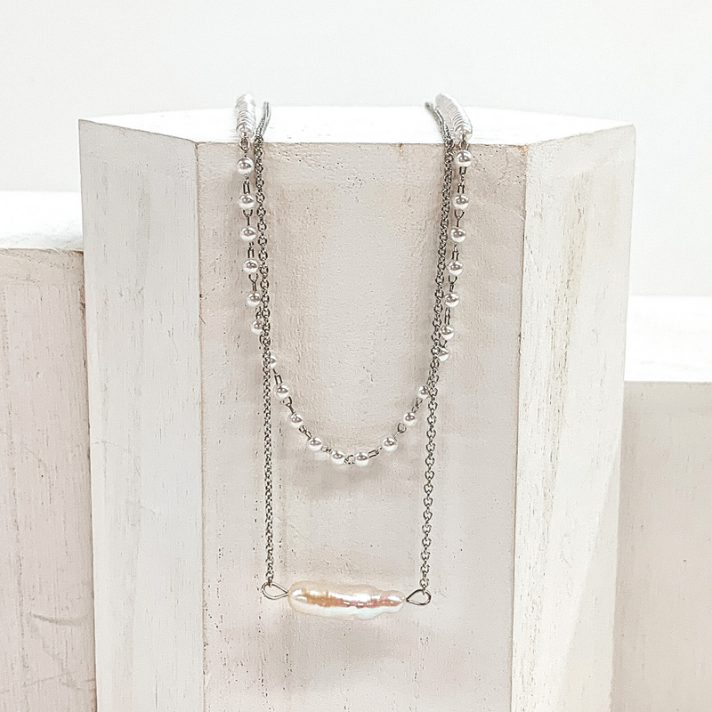 This is a silver, two stranded necklace that includes one silver chained necklace with an irregularly shaped pearl pedant and a pearl beaded necklace. This necklace is pictured laying on a white block on a white background.