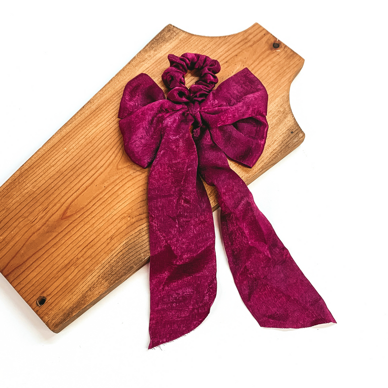 This is a maroon satin scrunchie with a long bow, this scrunchie is taken on  a brown necklace board and on a white background.