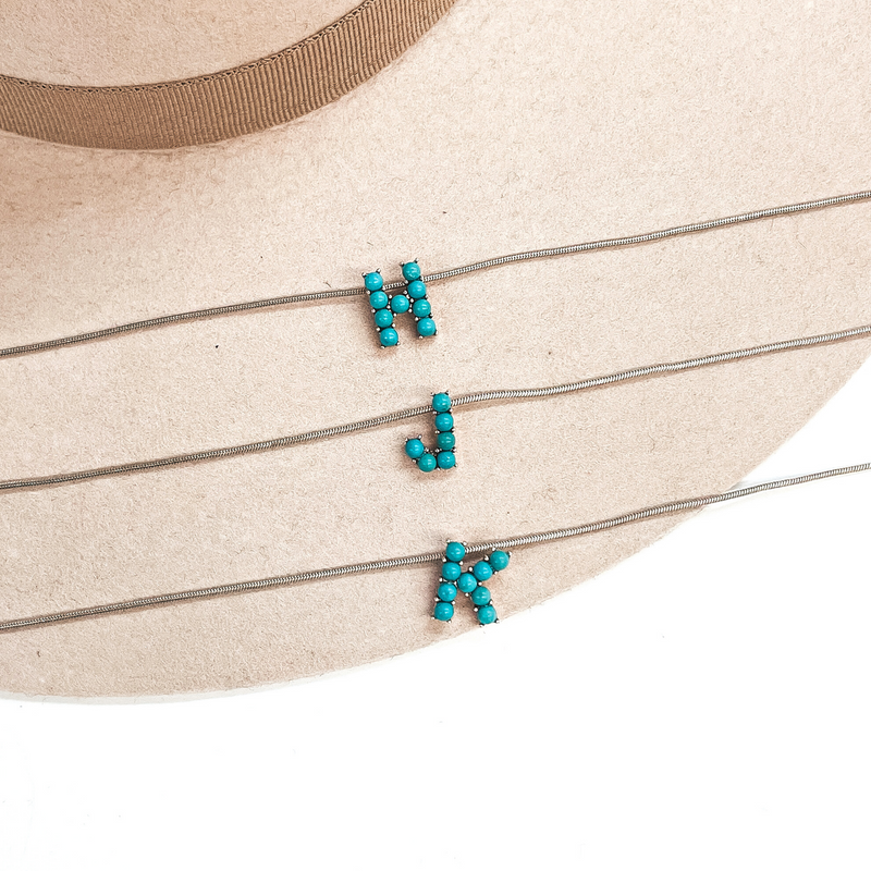 These are three turquoise stone initial necklaces with a thin, silver  snake chain. From top to bottom; H, J, K. These necklaces are taken  laying on a beige felt hat and on a white background.