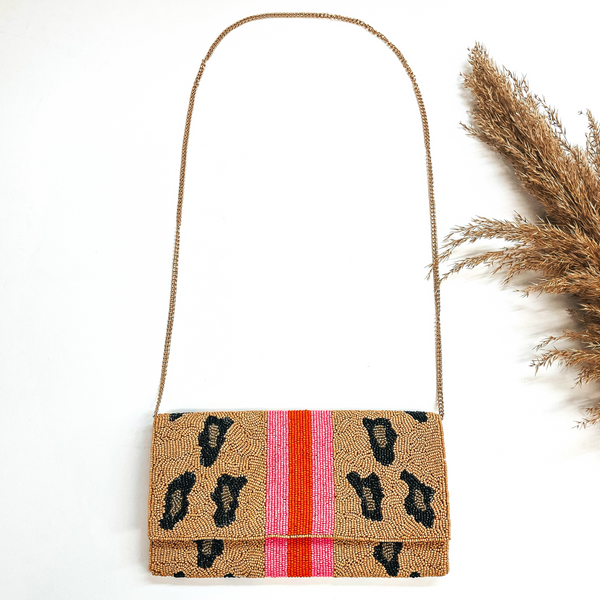 This is a seedbeaded clutch purse with a gold tone chain. This bag is in brown and  leopard print in black and gold. With  three stripes in the middle, two hot pink and one orange line in the middle. This bag  is taken laying on a white background with a brown plant in the side as decor.