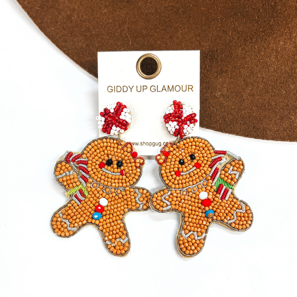 These are gingerbread seedbeaded earrings in tan. The stud part is white and red like a candy cane and the gingerbread girl is hold a candy cane as well. The gingerbread girl hassilver stitching in the arms and legs, there are three beads on the stomach as 'buttons' in white,red, and blue. All around there is a a tan and gold stitching. This pair of earrings are laying on a dark brown felt hat brim and on a white background.