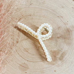 This is a long clear loop hair clip with pearls all around. This clip is laying on a slab of wood with a pink plant in the side as decor.