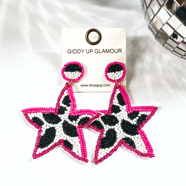 These are beaded star shape earrings in pink, black, and white. The center  is in black and white cow print, and pink beads all around. The post  back is half black and white, with pink beads all around as well. These  earrings are placed on an ivory thekitchenapproach earring card. These earrings  are taken on a white background with a disco ball in the side as decor.