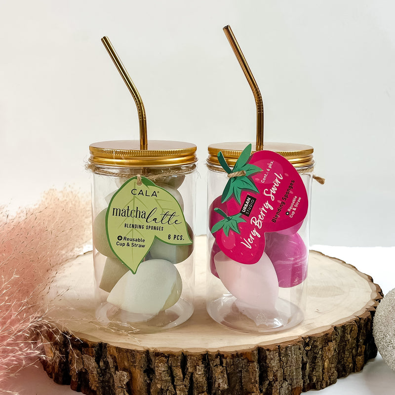 Two clear cups with gold lids and straws pictured on top of a piece of wood on a white background. Inside one cups is green colored sponges and the other cups has pink colored sponges. 