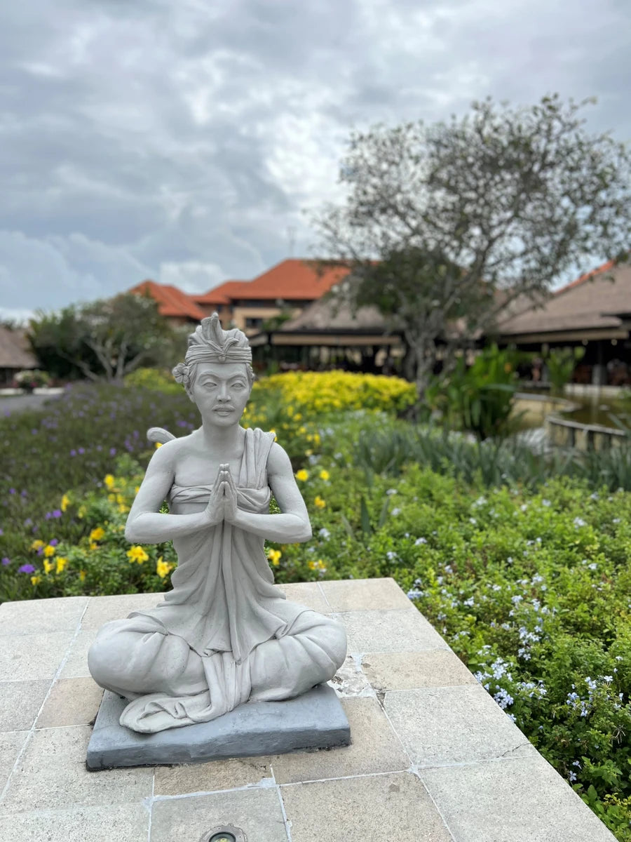 A statue of a Balinese woman sitting in prayer or meditation with a garden filled with flowers and a building in the background.