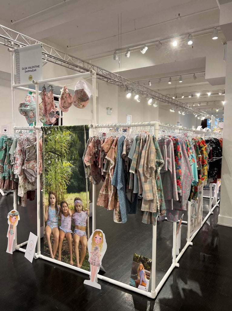 Olga Valentine’s display at the 2022 Playtime New York Tradeshow, showcasing their latest collection of girls’ fashions: swimsuits, swim tops and beach hats. Advertising material is also visible.