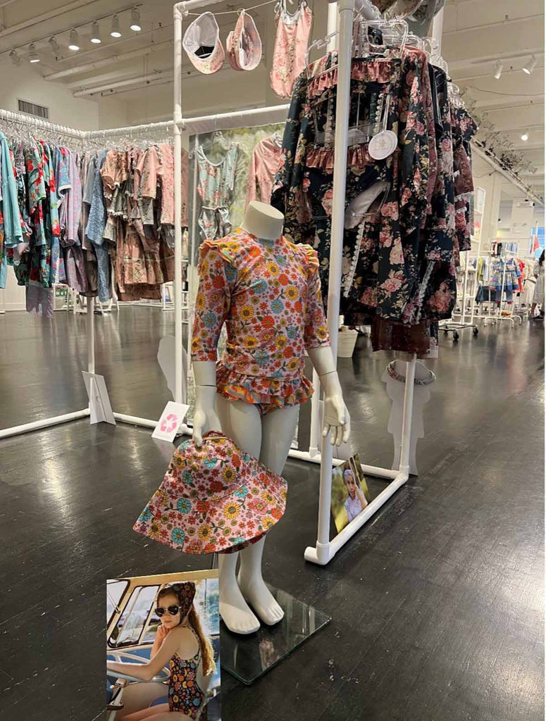 A 1:1 scale girls mannequin wearing the Olga Valentine Peace, Love & Happiness (Harmony) 2 Piece Rashi and Bottom Swim Set at the 2022 New York Playtime Tradeshow. The Mannequin is also holding the Peace, Love & Happiness (Harmony) Bucket Hat.