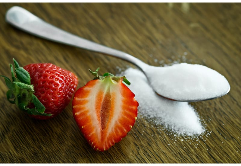 2 strawberries (one whole and one sliced) laid beside a teaspoon full of sugar
